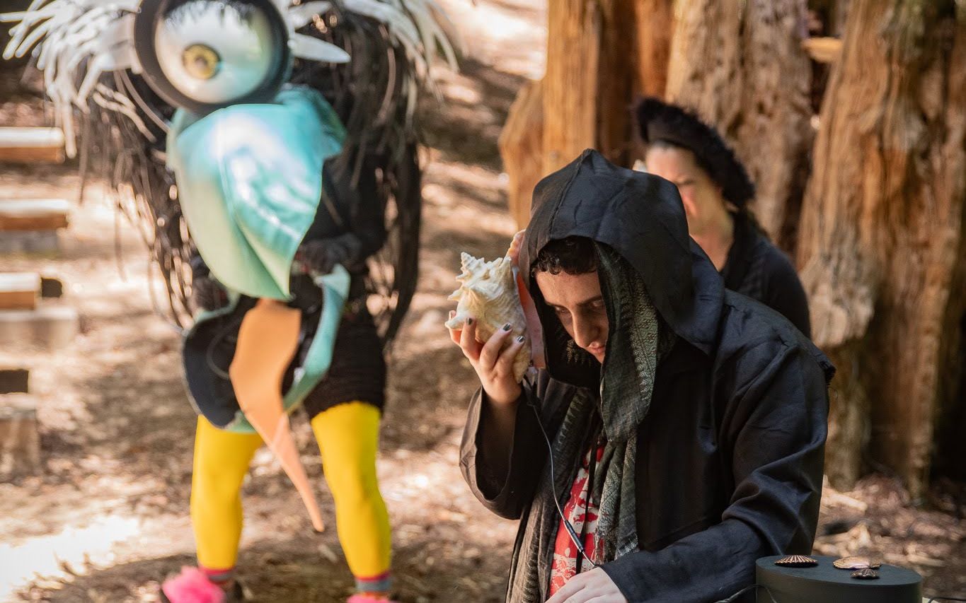 A hooded player holding a conch shell to their ear at the song-snippet music-puzzle device, beside a large feathered creature puppet at Adventure Camp.