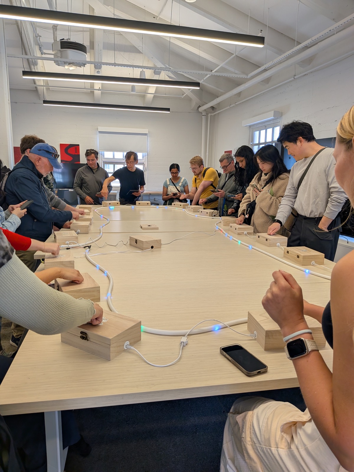 Players gathered around a large table knocking wooden stations linked by an LED-lit chase strip, playing Knock Band at the SF Pursuit scavenger hunt.