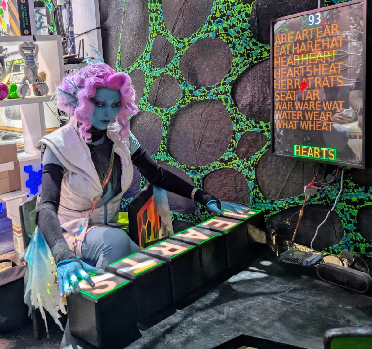 A costumed player spelling HEARTS on illuminated LEXACUBE letter cubes at Neotropolis, beside a live word-scoring display.